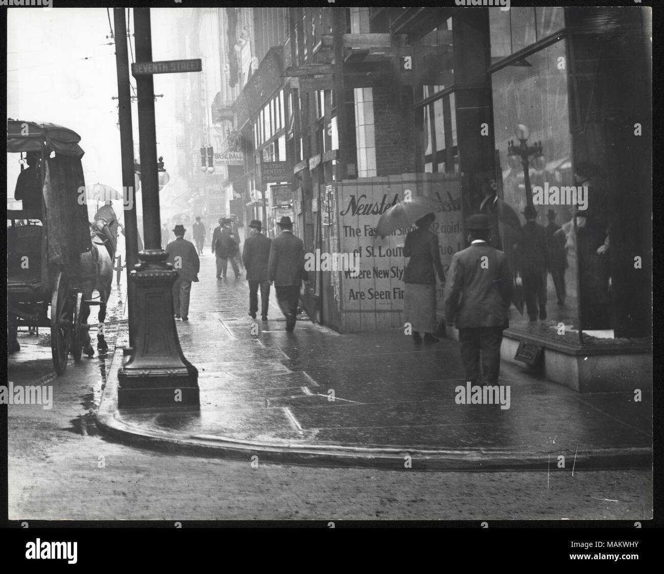 In orizzontale e di fotografia in bianco e nero dei pedoni camminare su Washington Avenue sotto la pioggia. La costruzione di edifici commerciali linea la pubblicità sul marciapiede per 'Baseball' e 'Dr. Dentista di pietra". Un carrello viene parcheggiato contro il marciapiede. Titolo: Washington guardando ad est dalla settima strada. [Dettaglio del 1-02-0357]. . Tra circa 1800 e circa 1900. Swekosky, William G., 1895-1964 Foto Stock