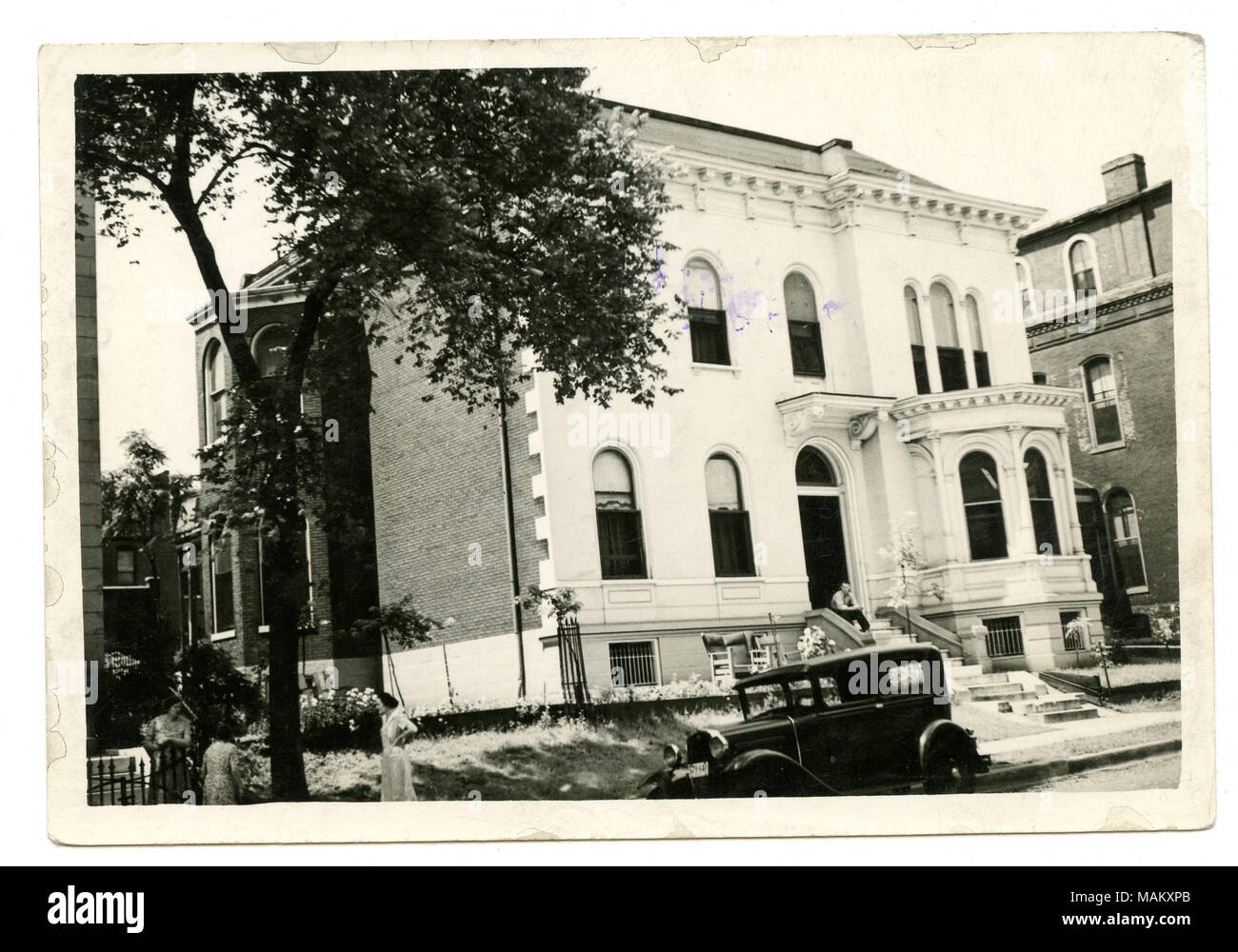 In orizzontale e di fotografia in bianco e nero di una di tre piani in pietra e mattoni con residenti cornicione e conci il rivestimento delle facciate laterali dell'edificio. Una finestra di baia con il cornicione è visibile sul lato destro della residenza accanto alla porta anteriore che è presente al di sotto modillions e una sporgenza. Il lato sinistro del residence dispone anche di una colonna di finestre a baia. Un uomo si siede sui gradini di fronte e tre donne sono visti nell'angolo inferiore sinistro della fotografia. Titolo: Wegler, Charles Residence. 1834 Kennett Place. . Tra circa 1878 e circa 1890. Swekosky, William G., 1895-1964 Foto Stock