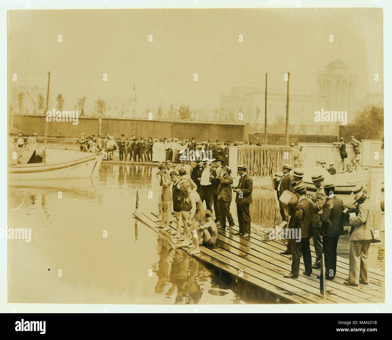 Fotografia orizzontale di nuotatori in piedi sul bordo di un dock. I giudici a stare dietro di loro. Vi è una piccola folla sulla riva e un treno dietro la folla. Edifici a partire dal 1904 della fiera del mondo può essere visto in background. Titolo: Avvio di un miglio di Handicap nuotare alle Olimpiadi del 1904. . 1904. Foto Stock