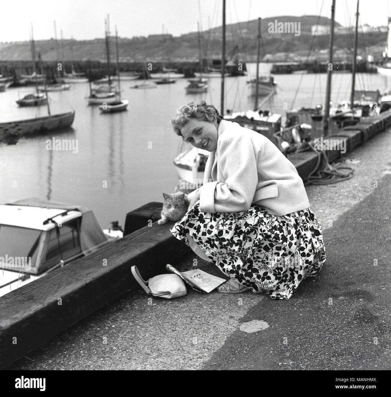 Degli anni Cinquanta, storico, un ben vestito lady in vacanza in ginocchio da un porto del bordo di accarezzare un gatto locale, England, Regno Unito Foto Stock