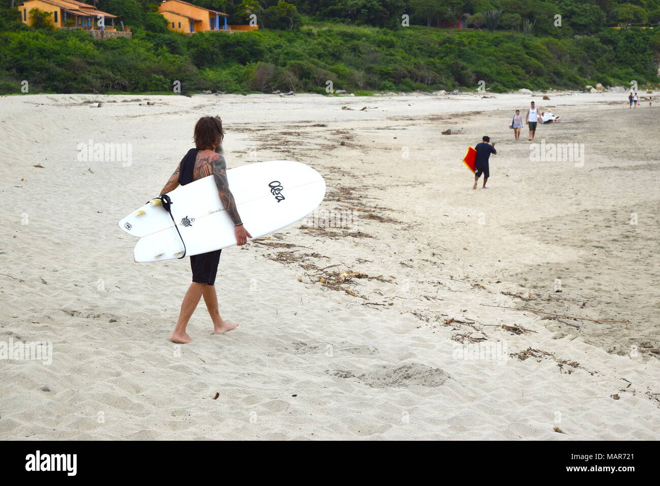 Surfer tatuato portante un white surf board passeggiate sulla spiaggia di sabbia in Messico Foto Stock