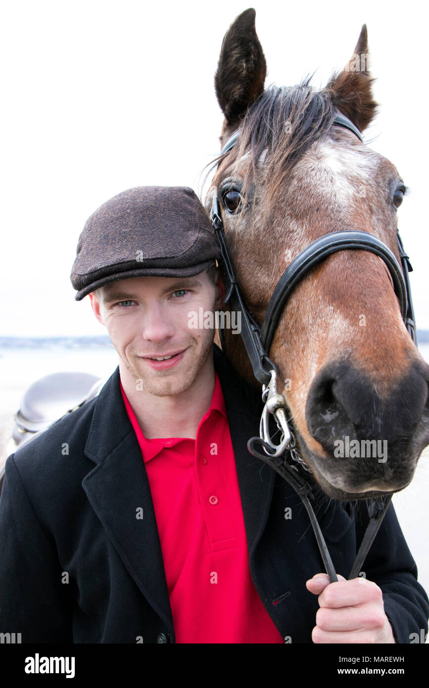 Uomo bello, pilota in piedi con il suo cavallo per ritratto. Indossando Maglietta polo rossa, flatcap e giacca nera sulla spiaggia. Foto Stock