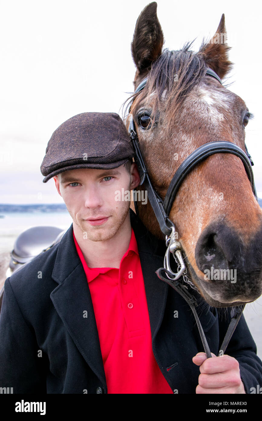 Uomo bello, pilota in piedi con il suo cavallo per ritratto. Indossando Maglietta polo rossa, flatcap e giacca nera sulla spiaggia. Foto Stock