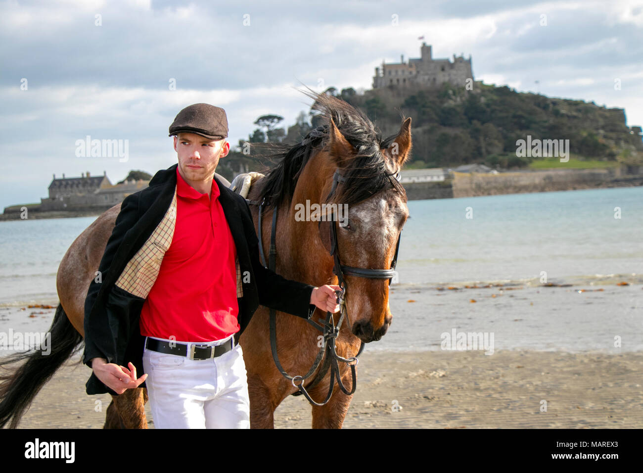 Bel cavallo maschio conducente in piedi camminando il suo cavallo, indossando il tappo piatto, pantaloni bianchi e stivali neri sulla spiaggia di fronte al St Michael's Mount Foto Stock