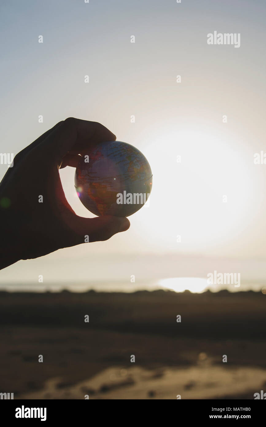 Primo piano di un giovane uomo caucasico con un globo mondo nella sua mano contro il cielo, in controluce, con il sole in background Foto Stock