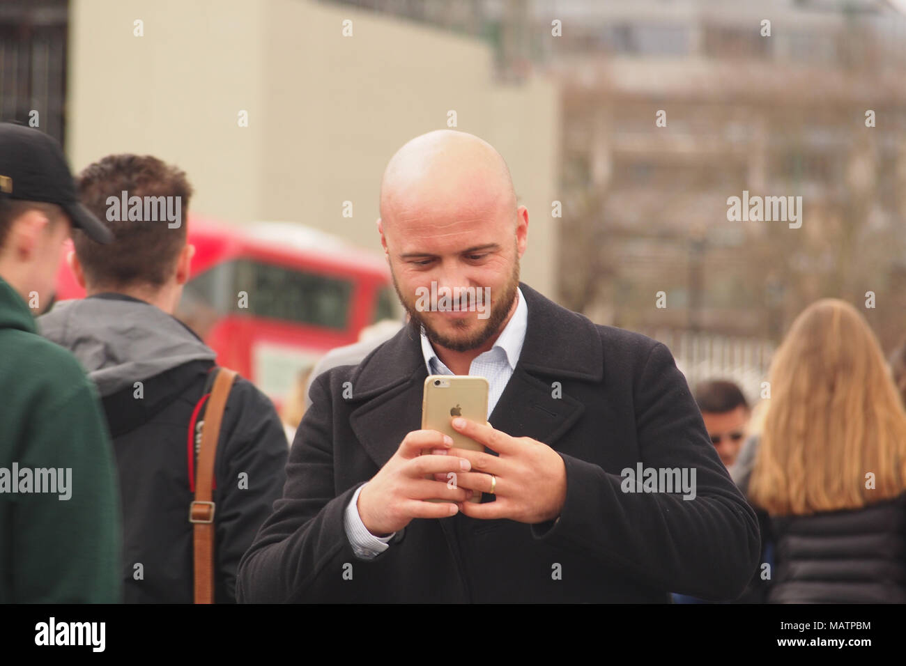 Un uomo che indossa un soprabito di prendere una fotografia sul suo smartphone a Londra tra una folla di pedoni e con un bus in background Foto Stock
