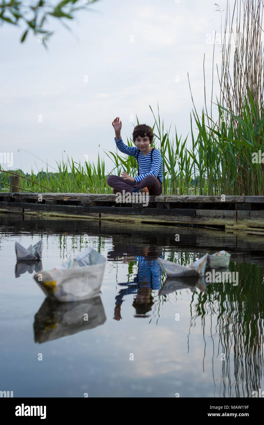 Un ragazzo su una banchina in legno osserva barche di carta. Foto Stock