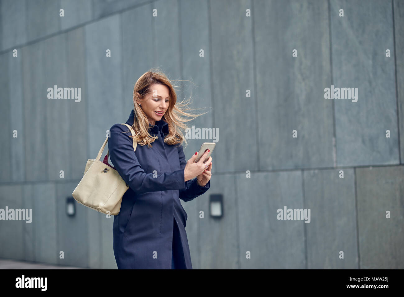 Di mezza età donna bionda passeggiate nel vento verso il basso una strada urbana a fianco di un muro grigio con i suoi capelli soffia nella brezza la lettura di un messaggio di testo su h Foto Stock