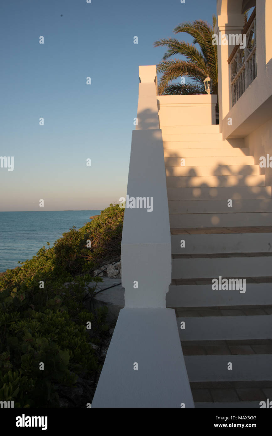 Volo di passi esterni su una lussuosa villa tropicali nei Caraibi che si affacciano sull'oceano al tramonto con un bagliore nel cielo all'orizzonte e fronde Foto Stock
