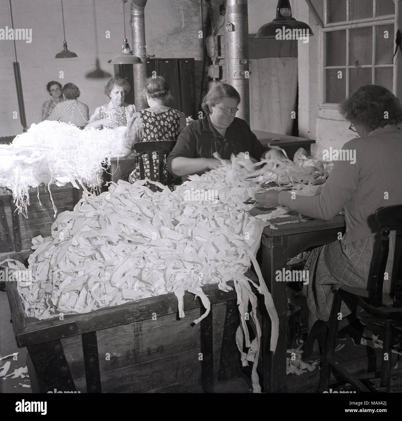Anni '50, storico, le donne che lavorano all'interno di un piccolo mulino di cotone seduta l'una di fronte all'altra a banchi che smistano grandi fasci di residui o off-tagli di materiale, forse cotone, Inghilterra, Regno Unito. Foto Stock