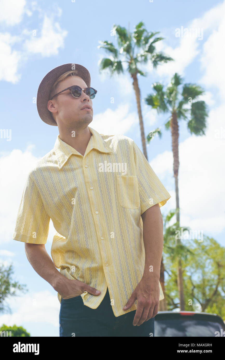 Un giovane uomo caucasico,indossando cappello, occhiali da sole e maglietta gialla, posa con palme e cielo blu in background.un maschio di vacanza il concetto di stile di vita Foto Stock