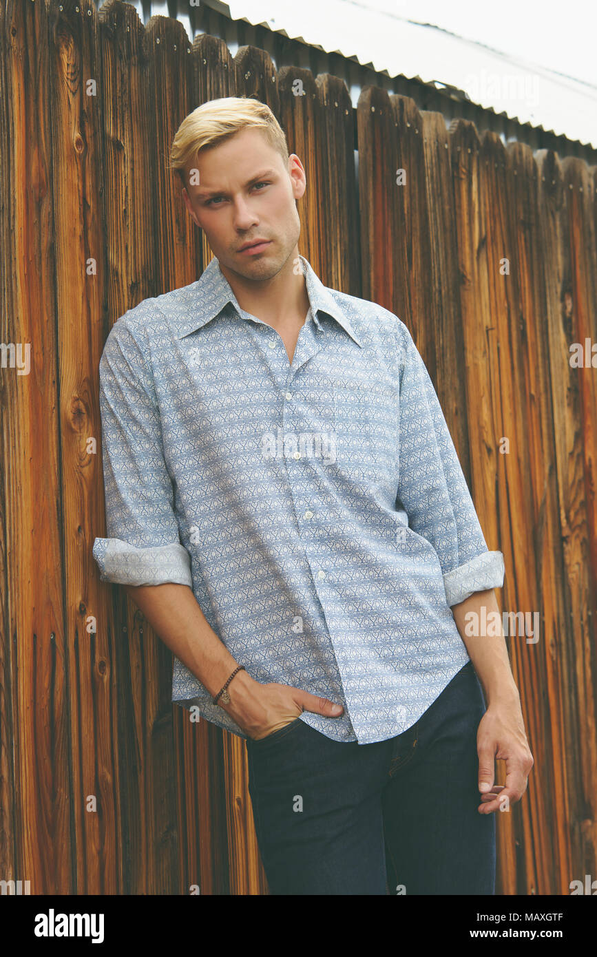 Una bionda uomo caucasico appoggiata contro una staccionata in legno, indossando uomini 70s magliette vintage, un uomo moda vintage concetto editoriale. Foto Stock