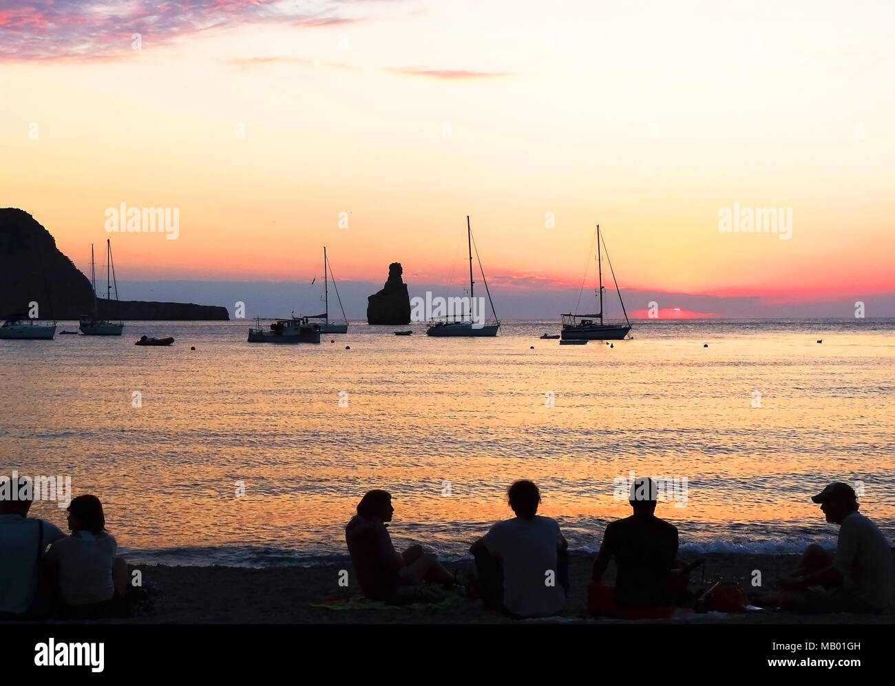 Tramonto di scena a Spiaggia Benirras, Isola di Ibiza. Formazione di roccia e sunbeam, vacanze estate scena. La gente a guardare il tramonto sulla spiaggia. Foto Stock