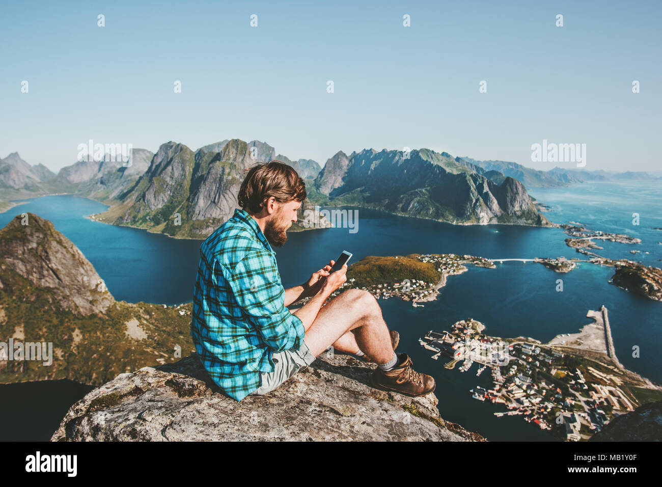 L'uomo traveler utilizza lo smartphone seduto sulla cima delle montagne in viaggio il concetto di stile di vita outdoor adventure Reinebringen mountain vista aerea Isole Lofoten Foto Stock
