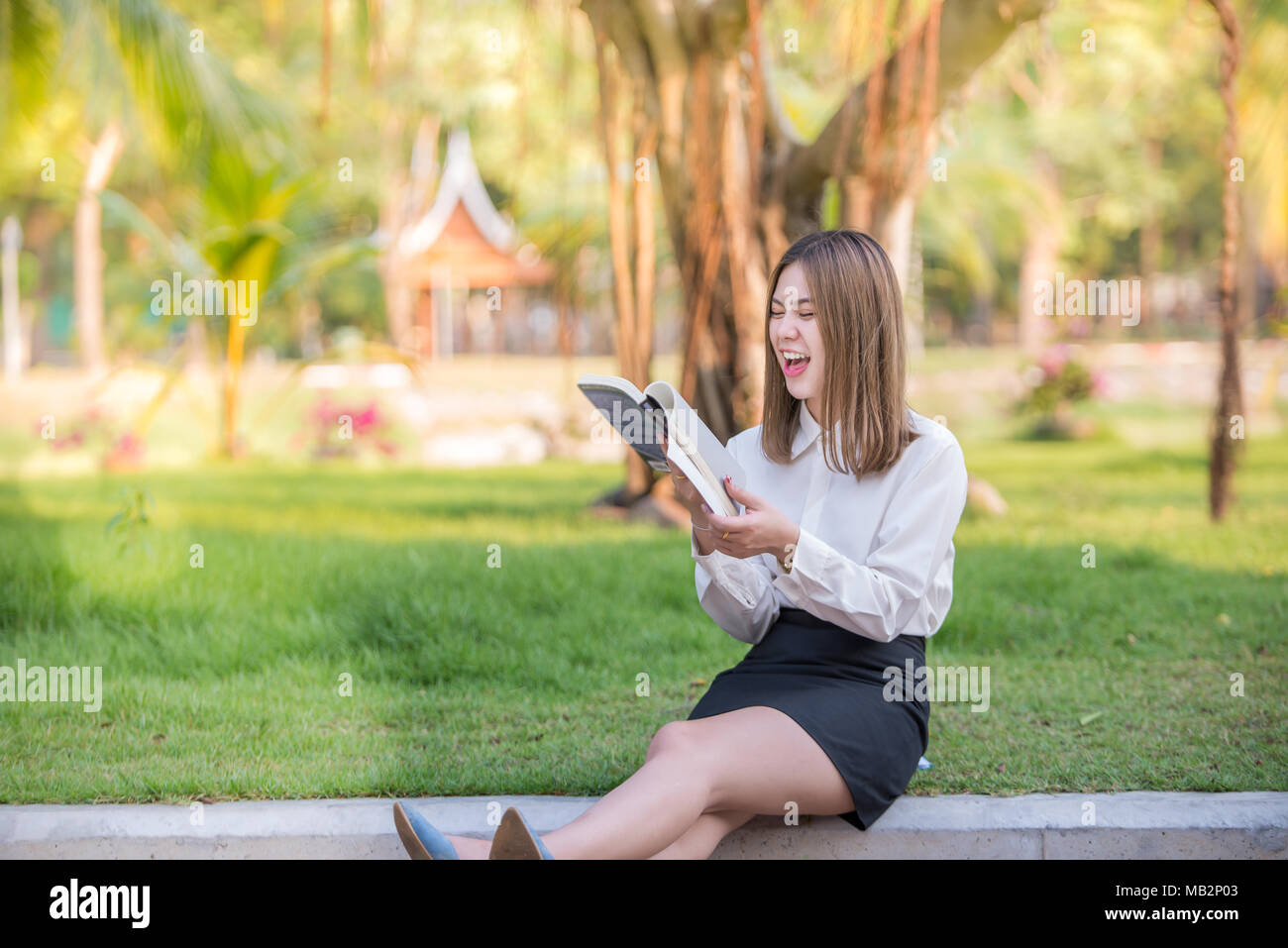 Happy business donna ridere e sorridere lei la lettura di un libro rivista in un parco urbano che indossa una minigonna Foto Stock