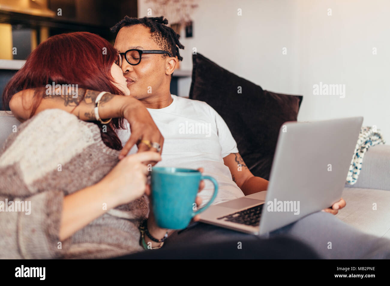 Giovani razza mista giovane seduto sul divano con il computer portatile e baciare. Coppia romantica trascorrere del tempo insieme a casa in soggiorno. Foto Stock