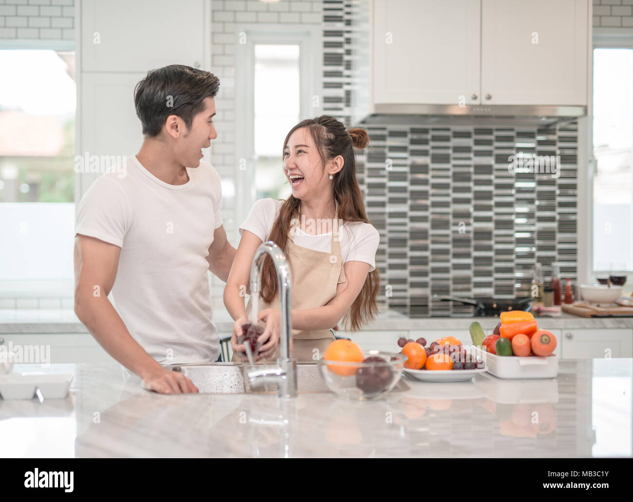 Felice giovane donna asiatica lavare frutta nel lavandino e uomo bello in piedi accanto a lei Foto Stock