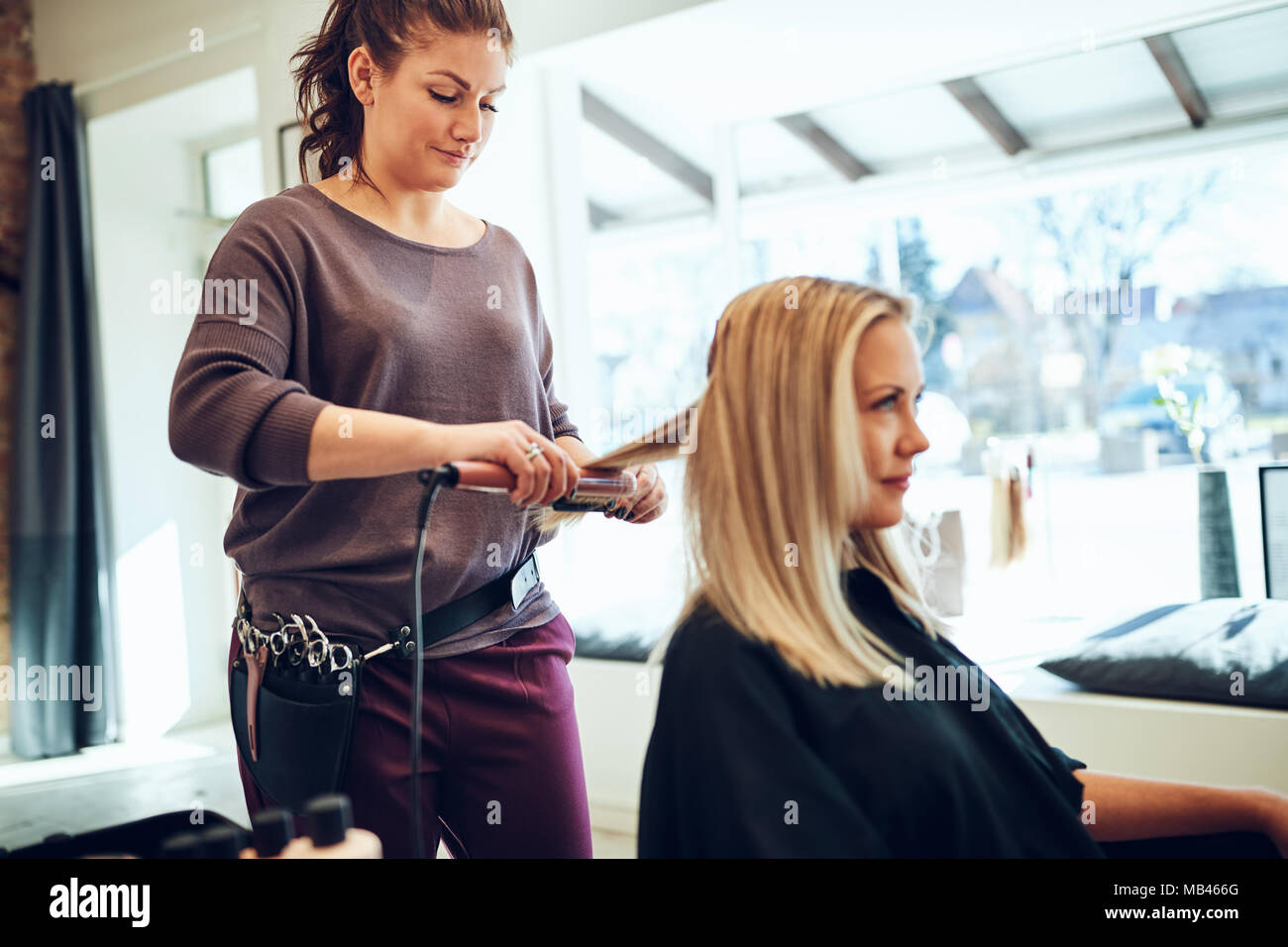 Sorridente bionda e giovane donna seduta su una sedia in un salone avente i suoi capelli raddrizzato dal suo parrucchiere Foto Stock