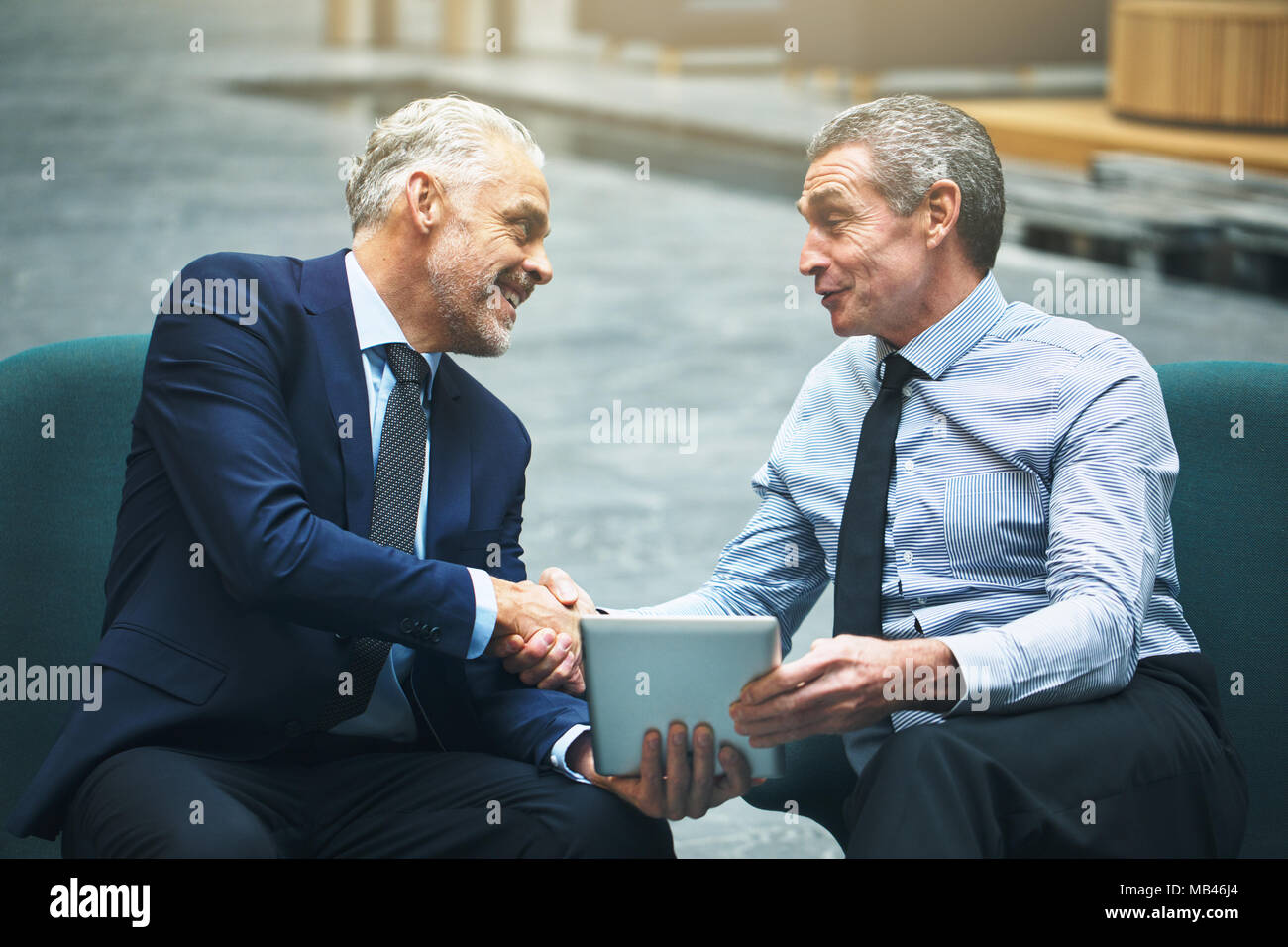 Due sorridenti imprenditori maturi stringono le mani insieme e utilizzando una tavoletta digitale seduti su sedie in un ufficio lobby Foto Stock