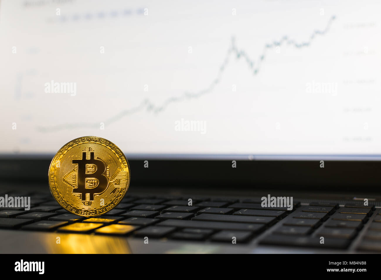 Bitcoin cryptocurrency coin in piedi su una tastiera portatile con ascendente grafico su sfondo sfocato Foto Stock