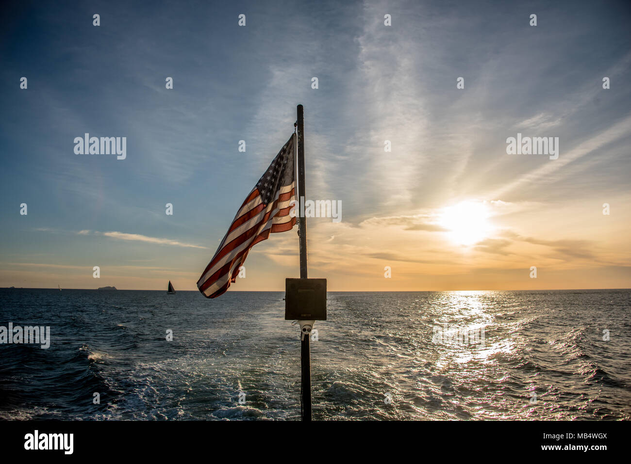 Bandiera americana in volo da una barca a vela in mare vista oltre la poppa contro un Cielo di tramonto con una scia in acqua dietro la barca Foto Stock