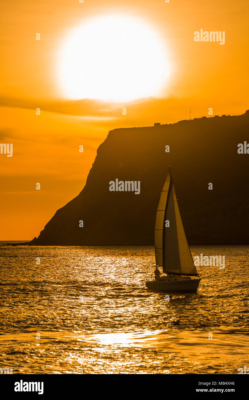 La vibrante arancione tramonto sull'oceano con una barca a vela vela passato un promontorio stagliano contro il cielo colorato e riflessi sull'acqua Foto Stock