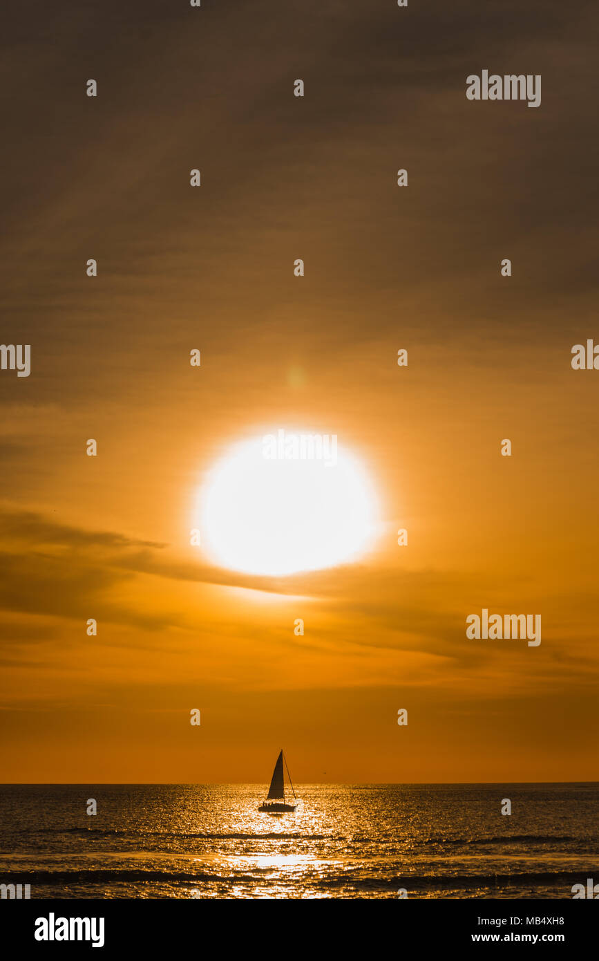 Lone Barca a Vela Barca a vela su un oceano tranquillo sotto un vivido arancione tramonto tropicale stagliano cast di riflessione del sole sull'acqua Foto Stock