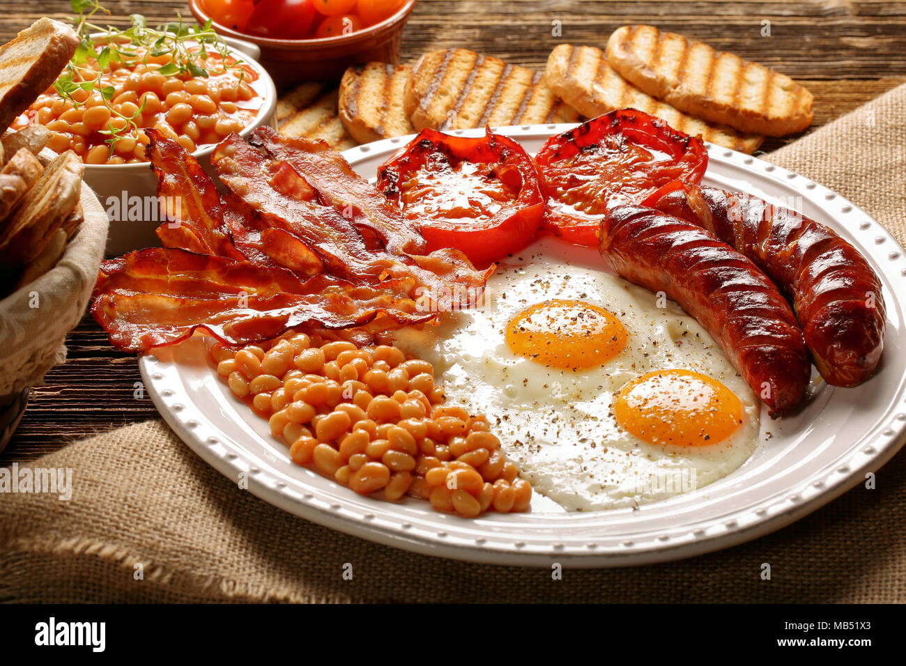 Prima colazione inglese con salsicce, pomodori grigliati, uova, pancetta, fagioli e pane sulla piastra bianca Foto Stock