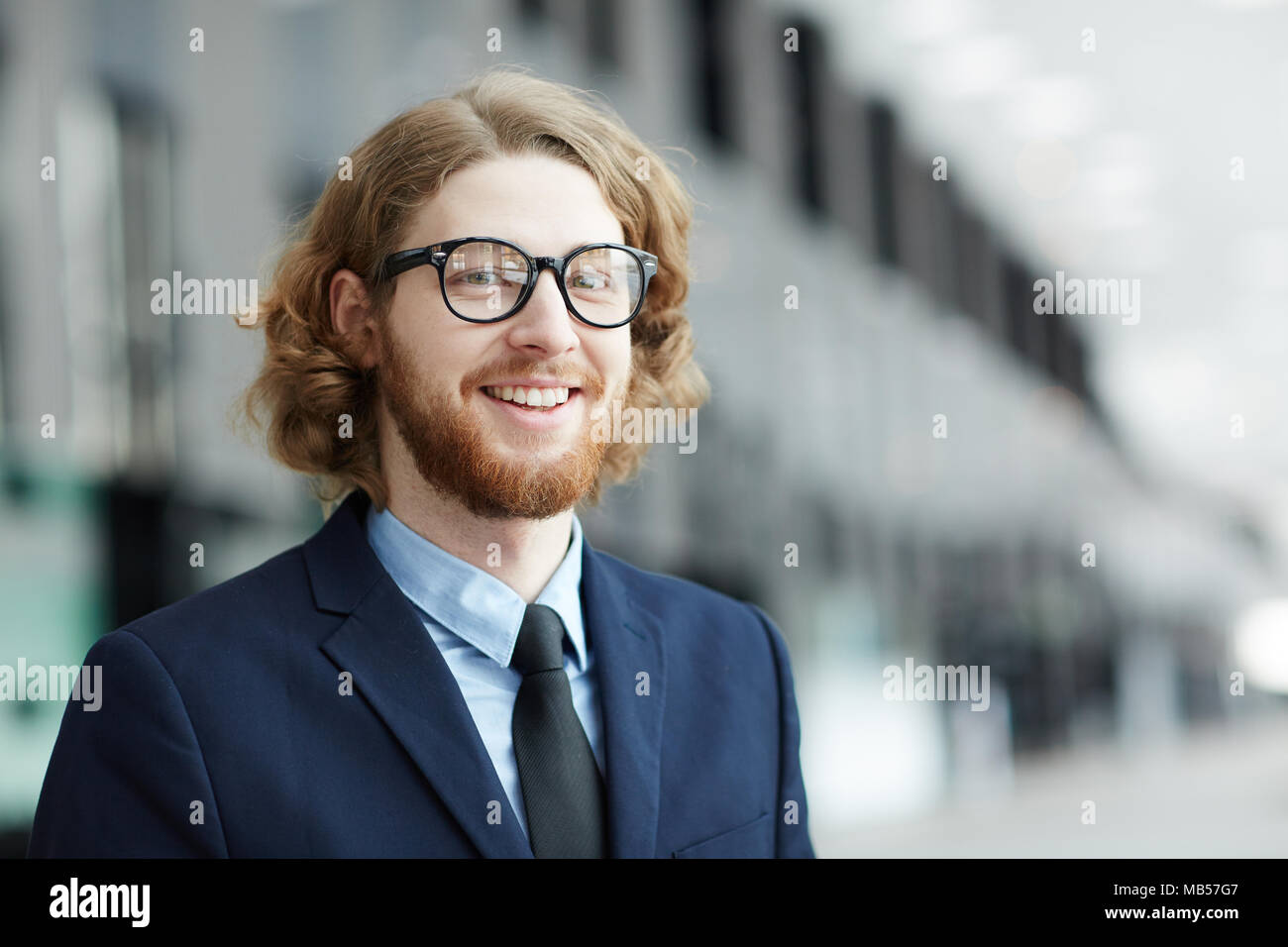 Giovani sorridente barbuto imprenditore nel convenzionale guardando la telecamera all'interno di edificio moderno Foto Stock