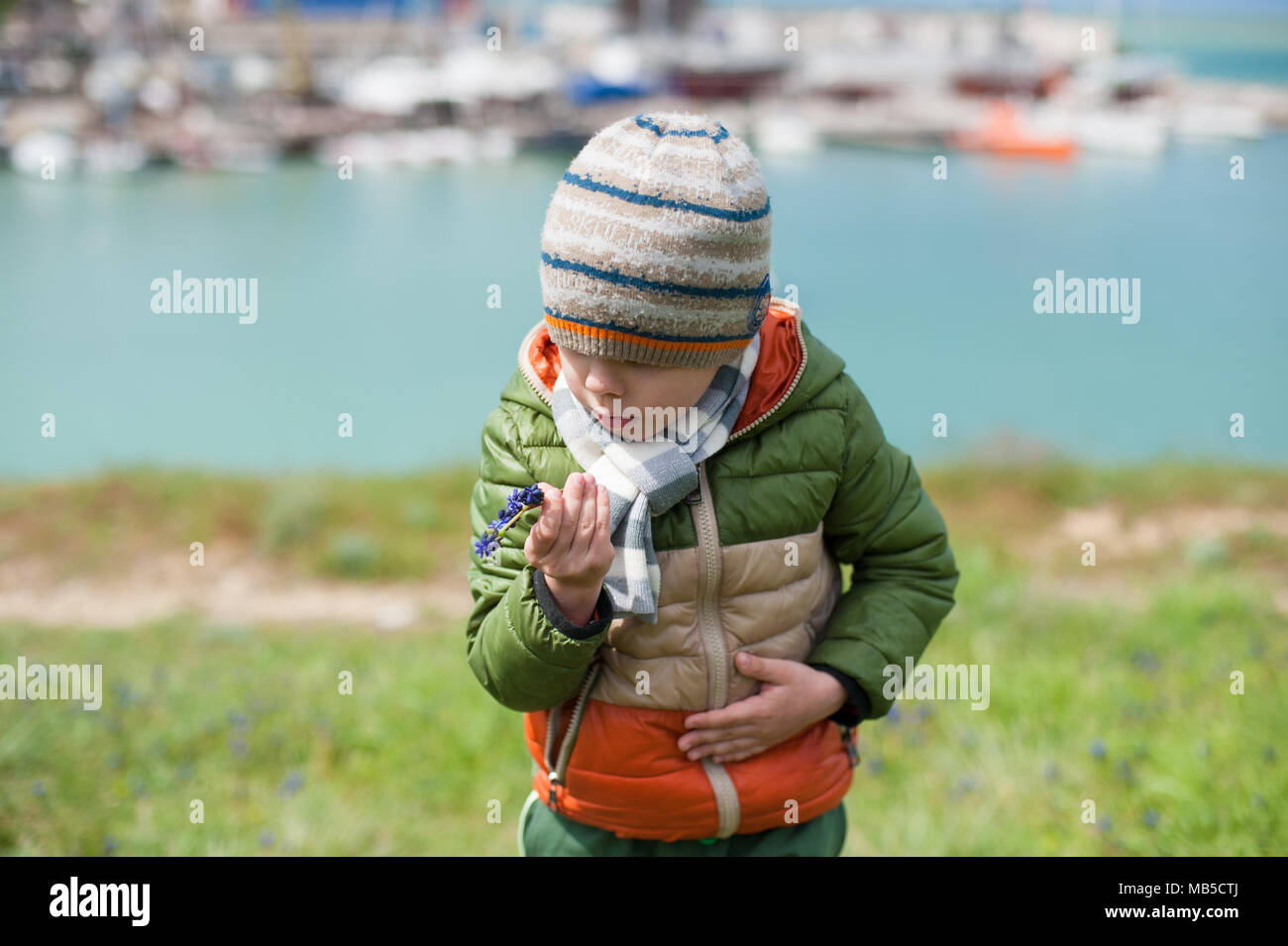 Curioso ragazzo in camicia, sciarpa e cappello sembra sorpreso per il Muscari fiore nel la sua mano sul mare sullo sfondo della baia in primavera Foto Stock