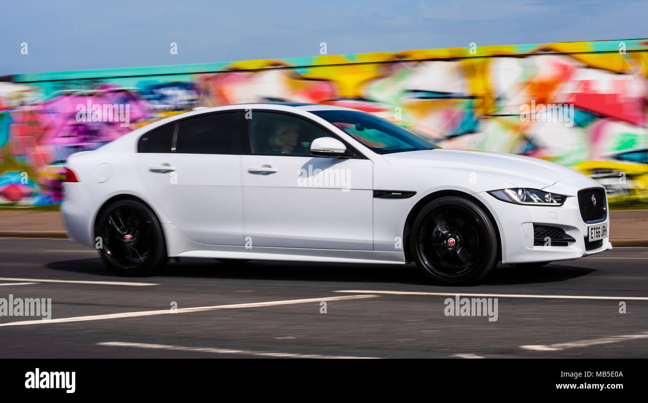 Jaguar XE 2016 automobile berlina che viaggiano a velocità passato coperto di graffiti in parete Southend on Sea, Essex. Veicolo bianco con sfondo colorato. Berlina Foto Stock