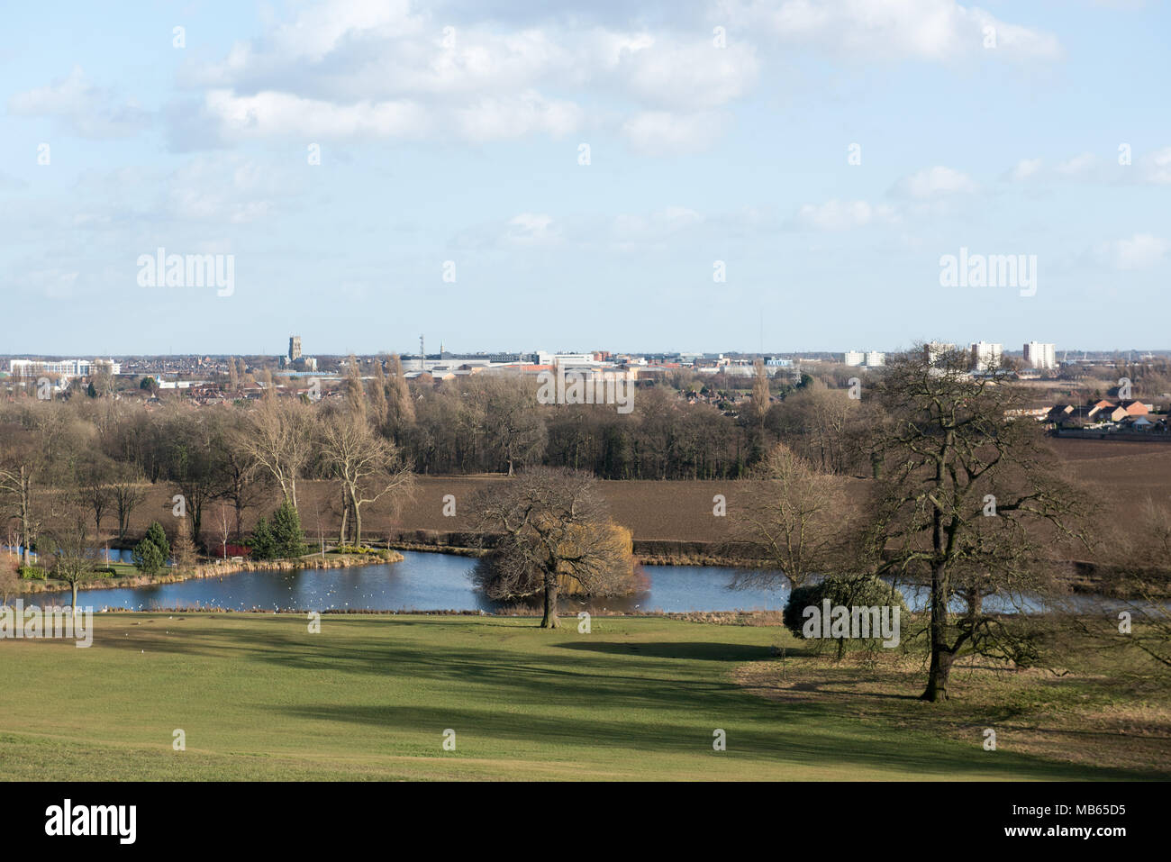 Un paesaggio tranquillo caratterizzato da un lago calmo, alberi sparsi e una vista panoramica di una città lontana sotto un cielo blu morbido con nuvole sparse Foto Stock