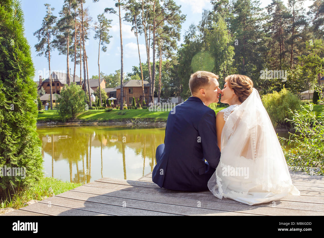 Sposi novelli kissing seduti sul molo in legno vicino al lago al giorno d'estate e di sole Foto Stock