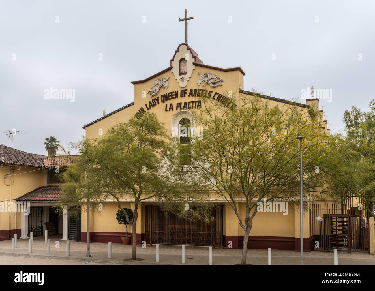 Los Angeles, CA, Stati Uniti d'America - Aprile 5, 2018: Giallo facciata frontale di piccole prima storica chiesa in città dietro una fila di alberi corti sotto il cielo d'argento. C grande Foto Stock