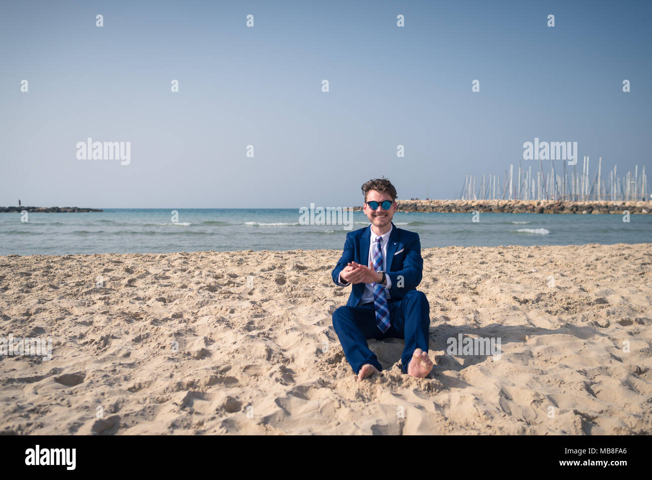 Insolito giovane in abito elegante si rilassa sulla spiaggia e sorridente. Funny turistico è seduto sulla sabbia e godersi la vacanza sullo sfondo del mare Foto Stock