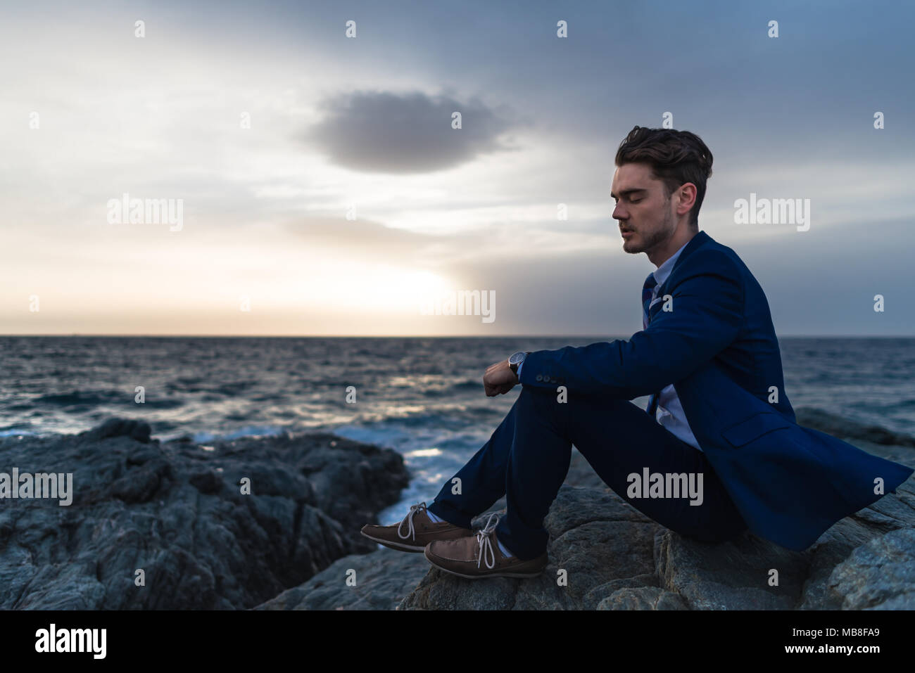 Stanco giovane elegante abito siede sullo sfondo del cielo e del mare. Malinconici guy meditando in prossimità dell'oceano di sera Foto Stock