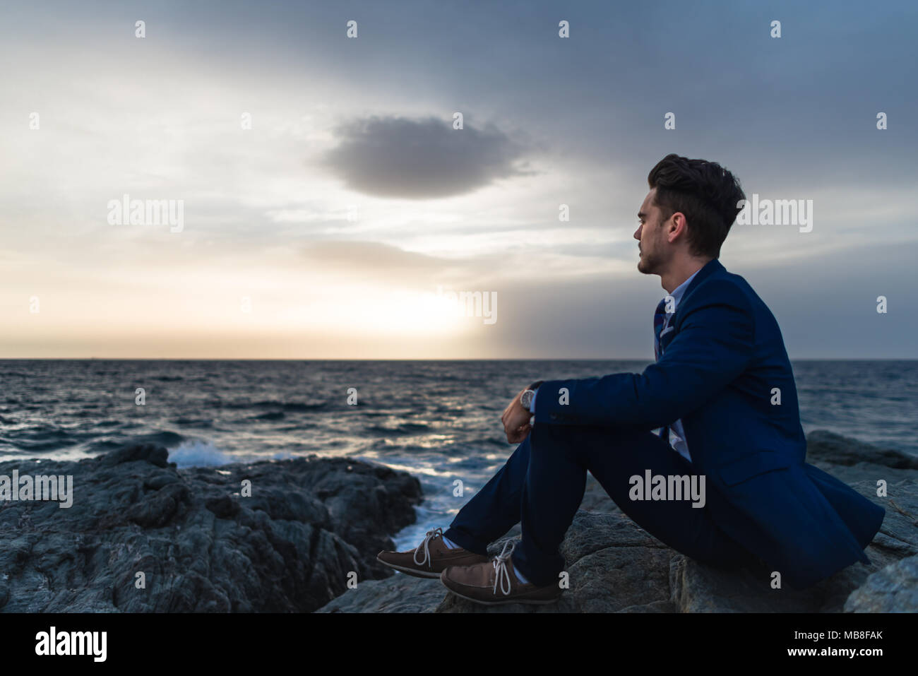 Giovane triste in elegante abito siede sullo sfondo del cielo e del mare. Malinconici guy meditando in prossimità dell'oceano di sera Foto Stock
