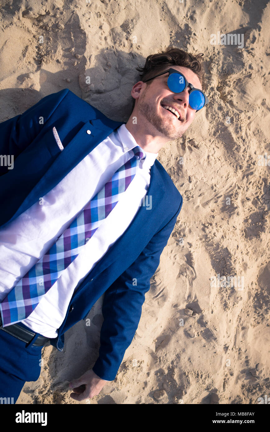 Insolito giovane in abito elegante si rilassa sulla spiaggia e sorridente. Funny tourist si trova sulla sabbia e godersi la vacanza Foto Stock