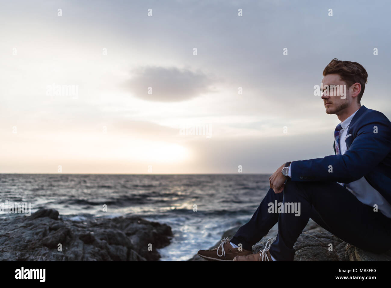 Giovane serio in elegante abito siede sulla costa vicino oceano. Giovane ragazzo pensando su sfondo di acqua e cielo Foto Stock