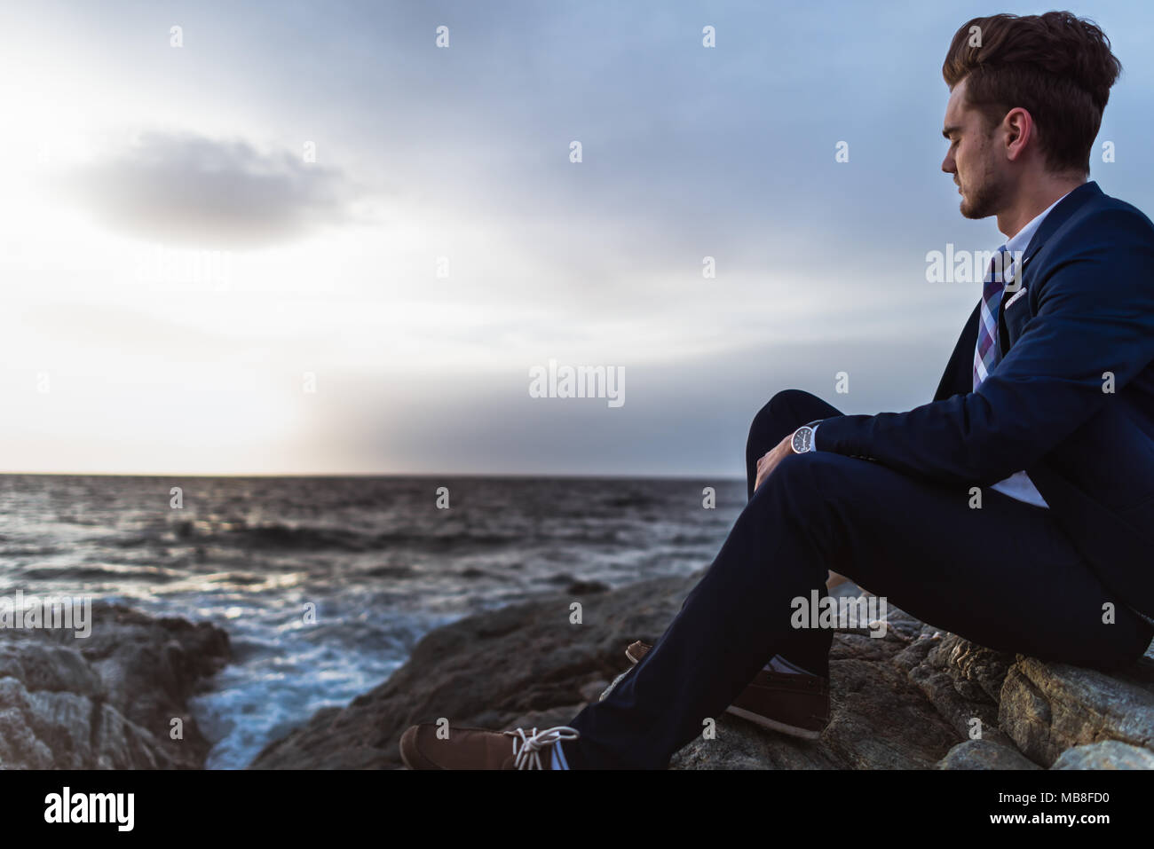 Malinconici uomo in tuta siede sulle rocce vicino al mare e di pensare. Giovane ragazzo meditando su sfondo del paesaggio marino e cielo Foto Stock