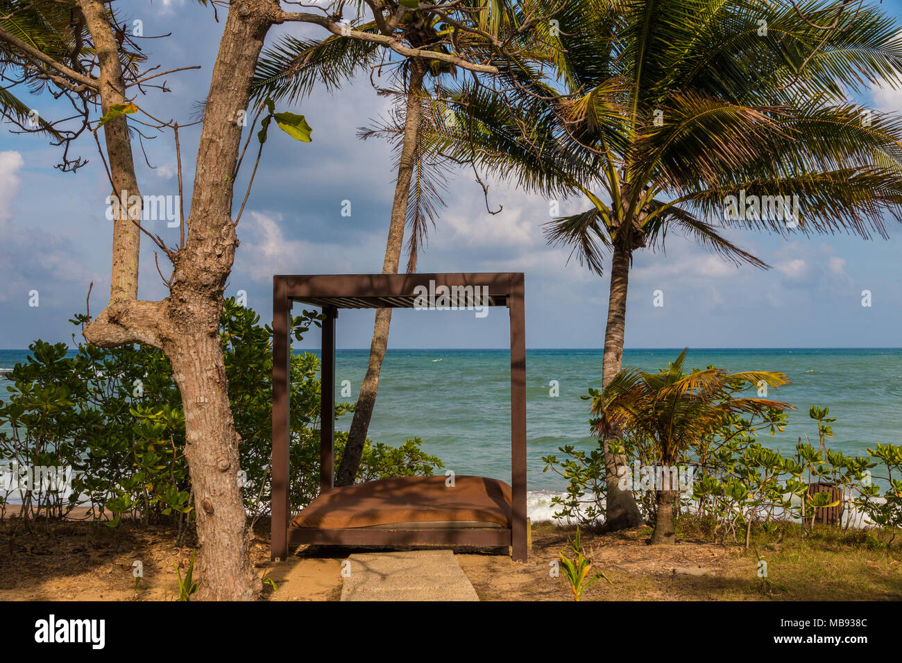 Un tipico scenario di vacanza, dove un open di legno cabana sulla spiaggia con un materasso all'interno è circondato da lussureggianti piante verdi e alberi di palma. Foto Stock