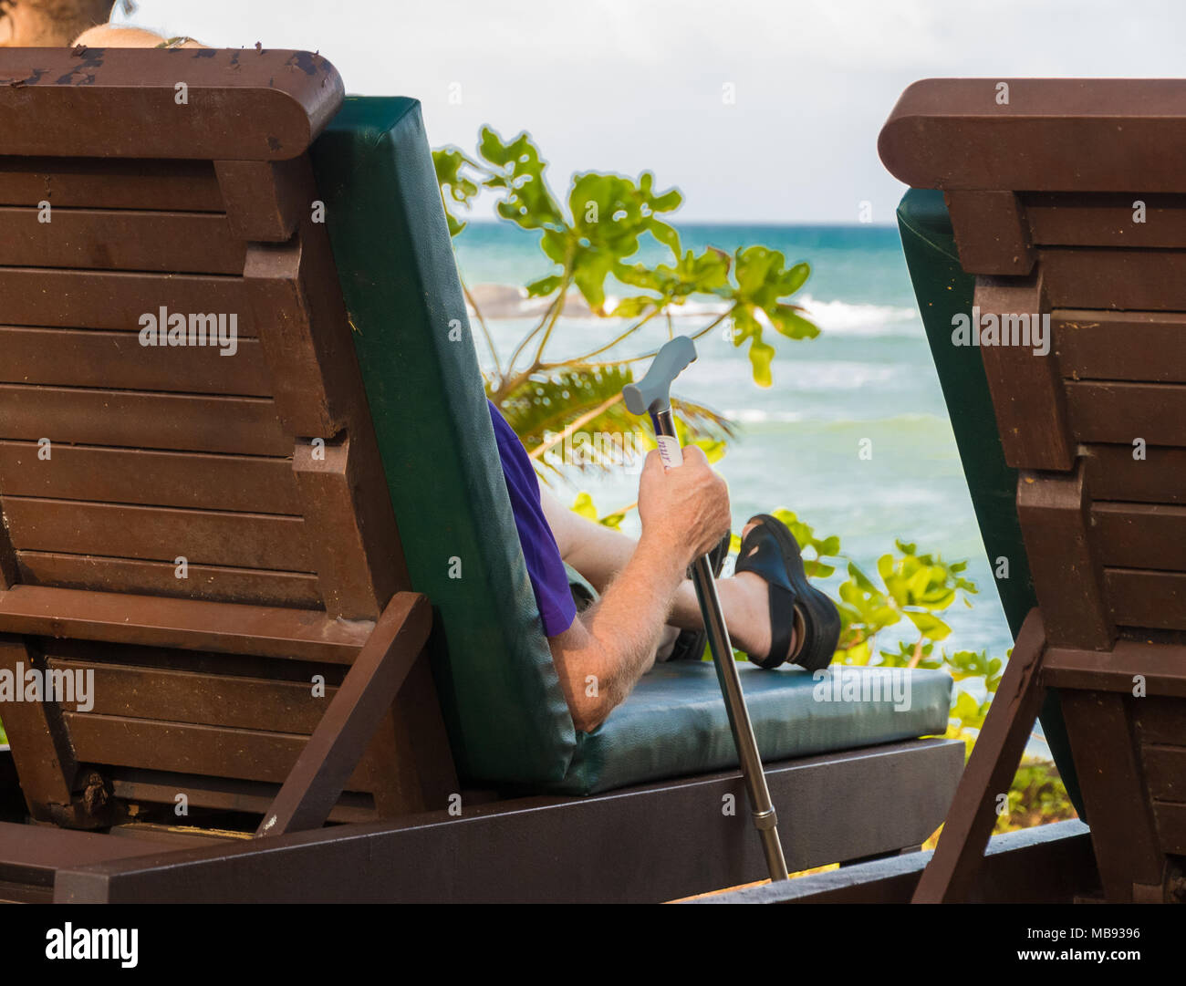 Un vecchio uomo occidentale con un bastone da passeggio, seduti su una sedia in spiaggia e godersi il suo pensionamento all'estero. Presa nel febbraio 2018 in Terengganu, Malaysia. Foto Stock