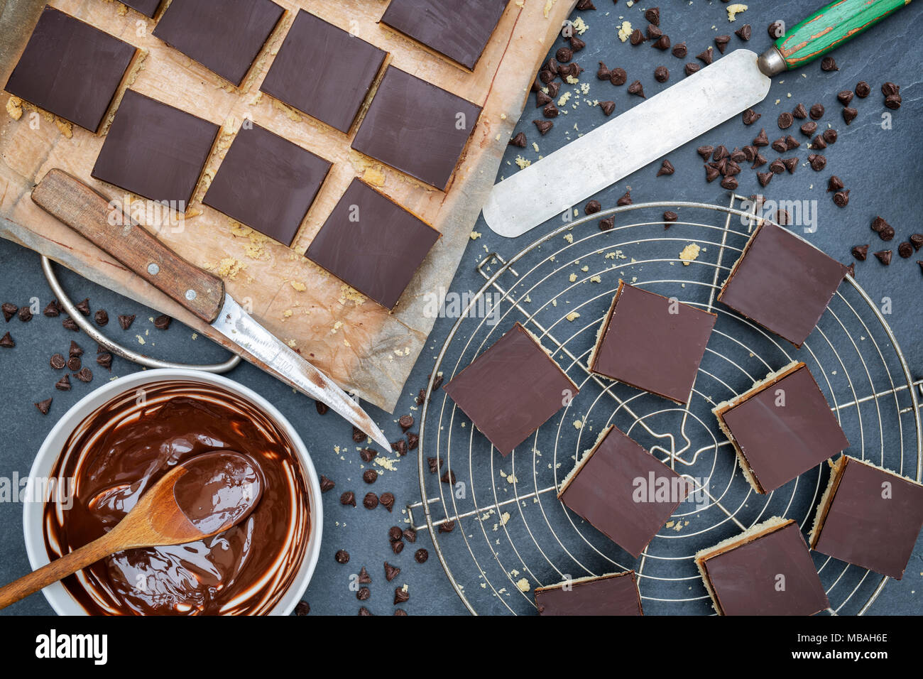 Caramello / biscotti frollini milionari piazze con un pallet il coltello e il cioccolato fondente fuso in un recipiente su un sfondo di ardesia Foto Stock