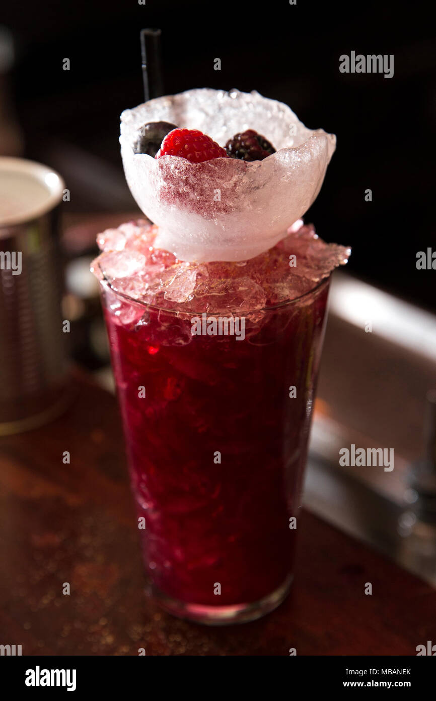 Colpo verticale di un colore rosso intenso cocktail su un banco di bar con un bicchiere di ghiaccio e bacche di esso come decorazione.Cocktail è stata fatta con frutta organica Foto Stock