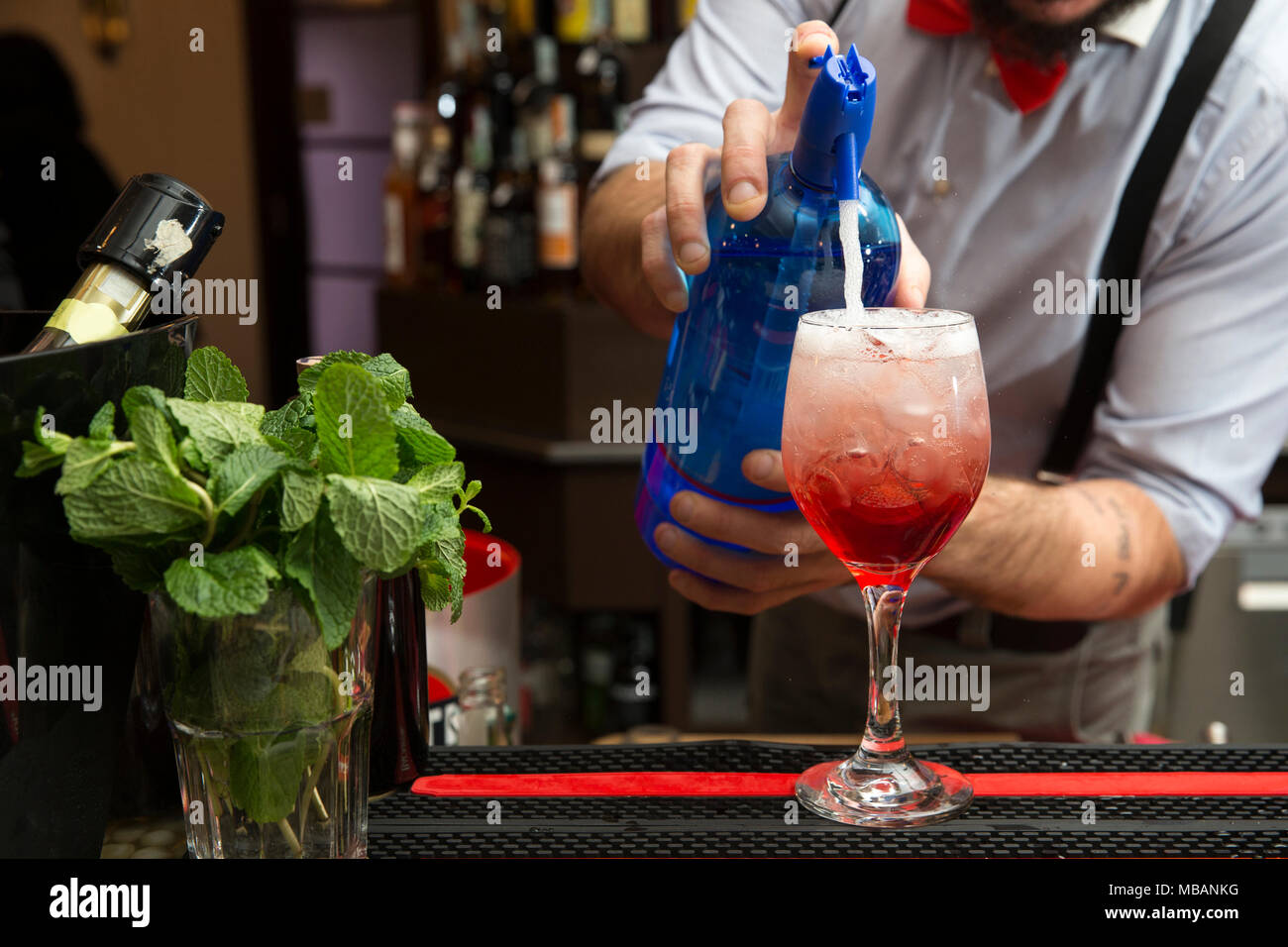Primo piano di un barman con un rosso papillon, rendendo un rosso spritz su una barra couter. Egli versando acqua di soda nel bicchiere pieno di ghiaccio. Foto Stock