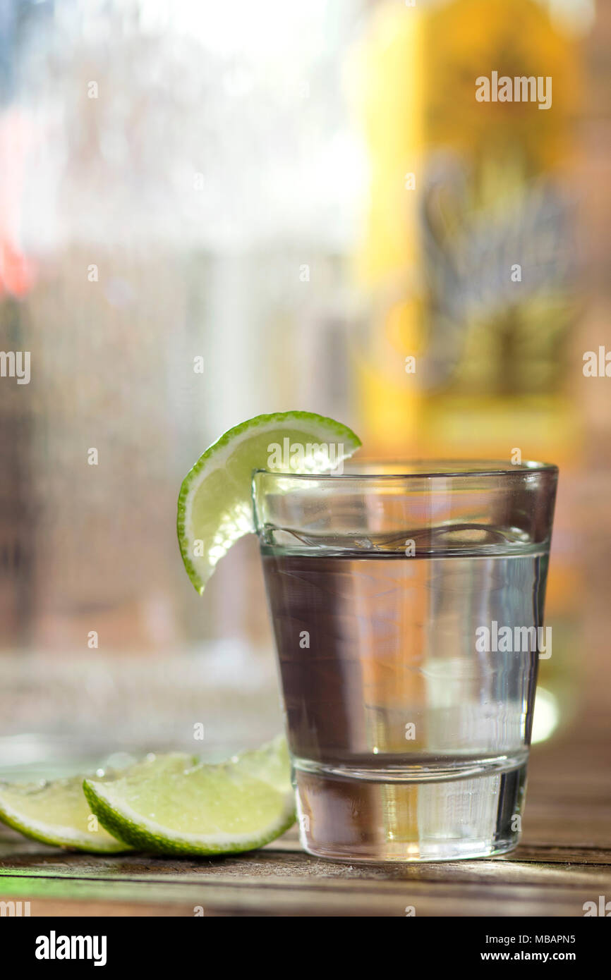 L'alcol colpo di vetro, tequila o vodka, con un lime / fetta di limone, closeup, con molte bottiglie in background, al di fuori della messa a fuoco Foto Stock