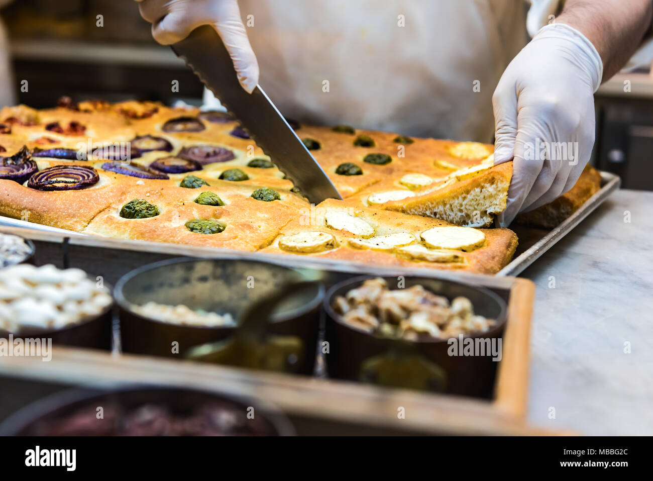 Il taglio foccacia pane con coltello, noci, spezie in contenitori e condimenti cotto in forno, lo chef del ristorante cucina con igiene sanitaria guanti Foto Stock