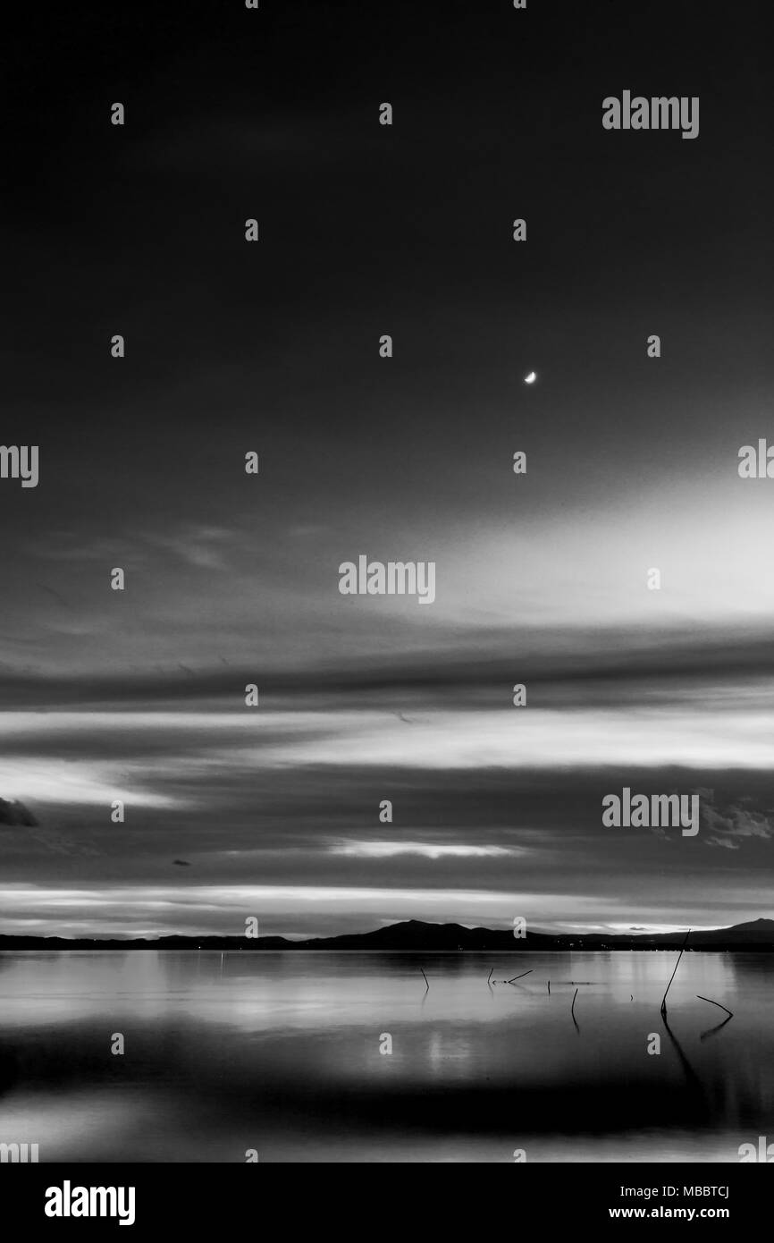 Bellissima vista del lago Trasimeno (Umbria, Italia) al tramonto, con toni del bianco e del nero e la luna nel cielo Foto Stock