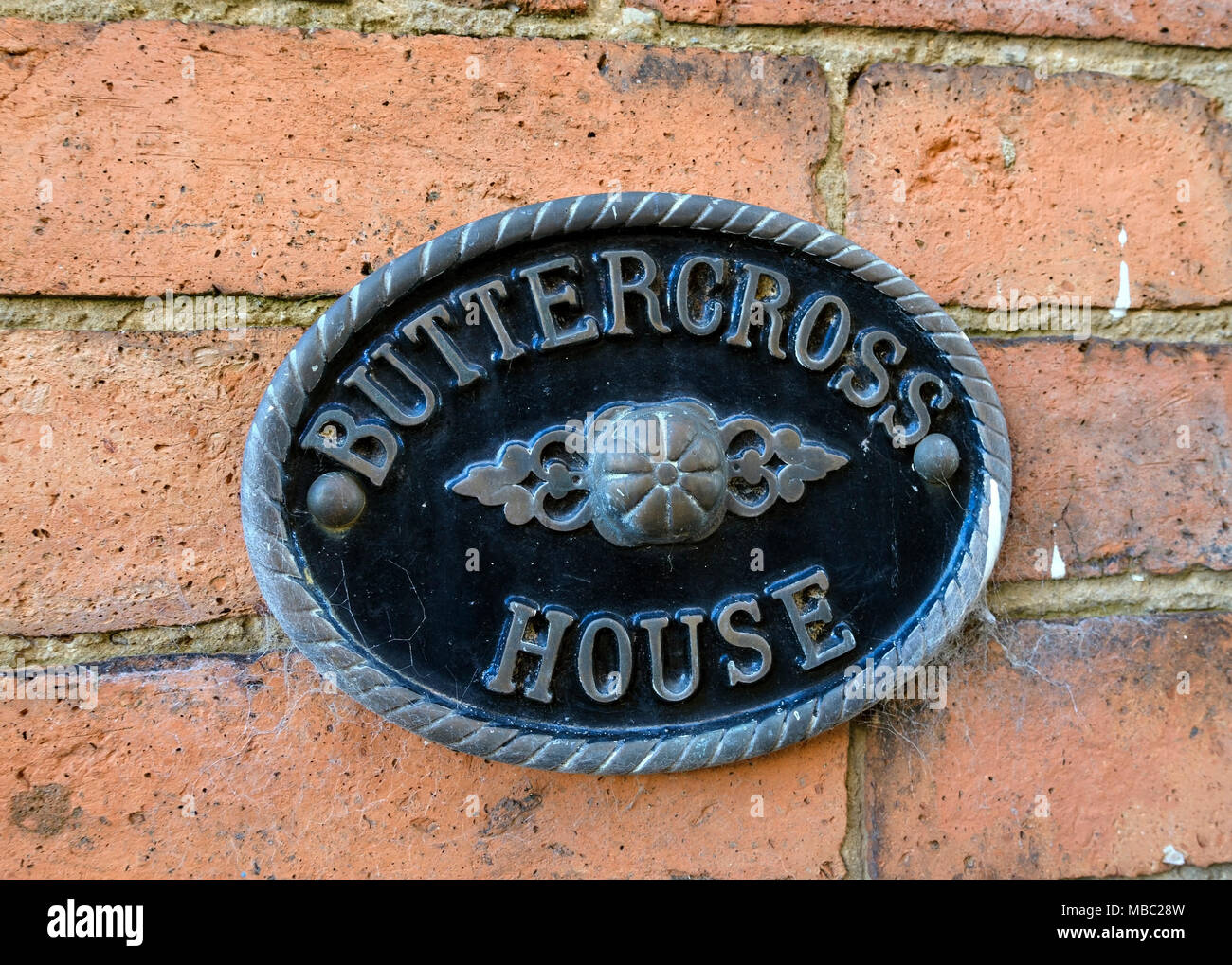 Smart ornato di forma ovale in metallo fuso segno casa per casa Buttercross montato sul vecchio rosso muro di mattoni, Oakham, Rutland, Leicestershire, England, Regno Unito Foto Stock