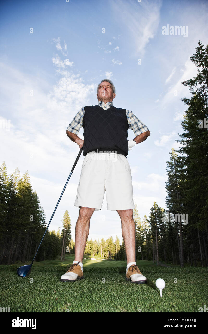 Un uomo in piedi le mani sui fianchi con un club di golf in mano bassa angleview, sul campo da golf, alberi di pino e il paesaggio. Foto Stock